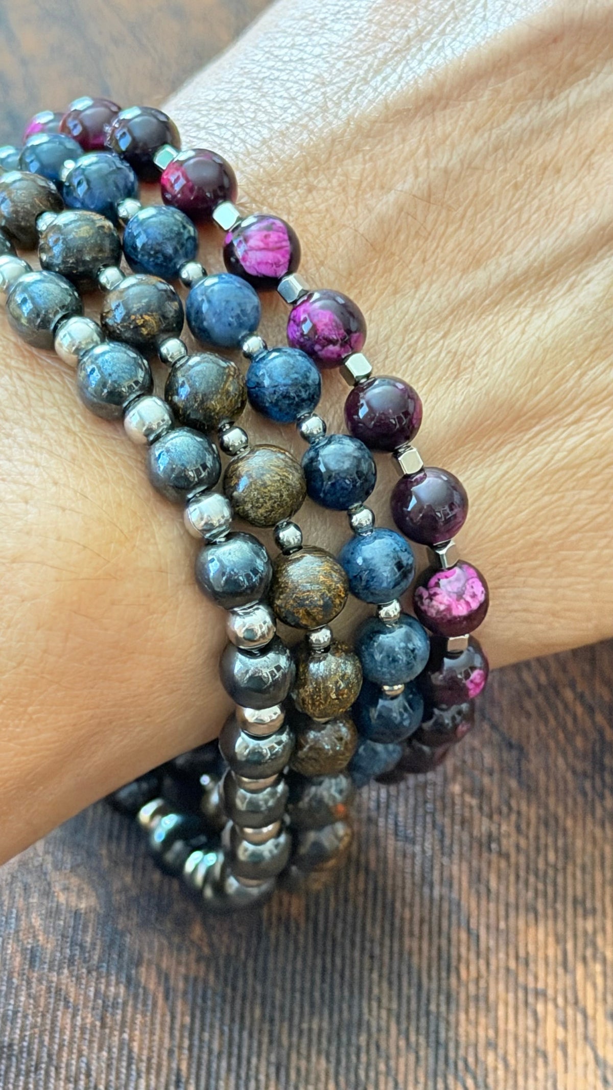 A close-up of a person's wrist wearing multiple beaded stretch bracelets. The bracelets feature polished round beads in deep blue, smoky gray-brown, and purple with metallic silver spacers, set against a dark wood background.